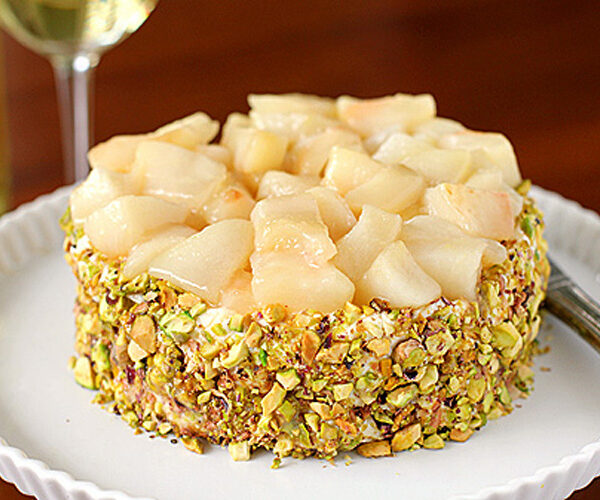A round dessert topped with diced pears, coated on the sides with chopped pistachios, sits on a white plate. A glass of white wine is visible in the background.