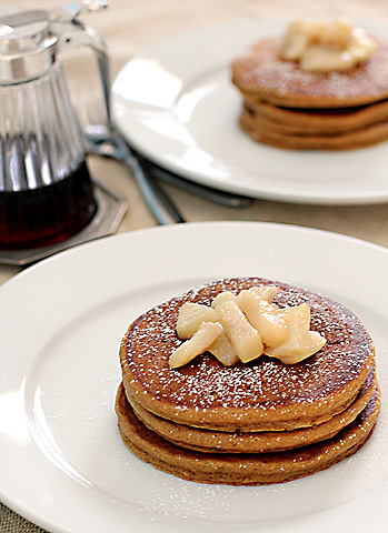 Three stacked pancakes topped with chopped bananas and a dusting of powdered sugar on a white plate, with a syrup pitcher and another plate of pancakes in the background.