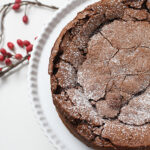 A chocolate cake with a cracked top dusted with powdered sugar sits on a white scalloped cake stand. Red berries and twigs are arranged on the white surface beside it.