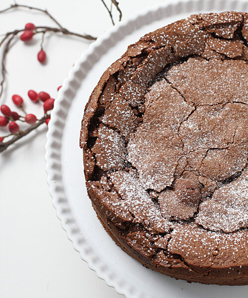 A chocolate cake with a cracked top dusted with powdered sugar sits on a white scalloped cake stand. Red berries and twigs are arranged on the white surface beside it.