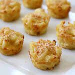 Bite-sized baked macaroni and cheese cups are arranged on a white plate, with a golden brown, crispy topping visible on each portion.