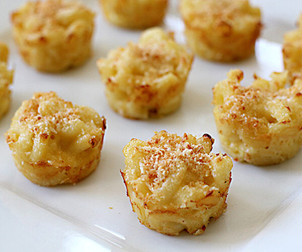 Bite-sized baked macaroni and cheese cups are arranged on a white plate, with a golden brown, crispy topping visible on each portion.