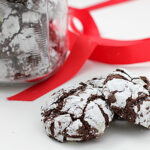 Two chocolate crinkle cookies coated in powdered sugar sit in front of a glass jar filled with more cookies, with a bright red ribbon in the background on a white surface.