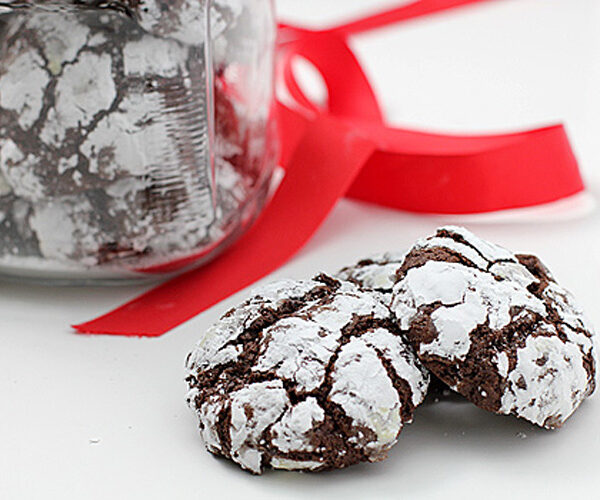 Two chocolate crinkle cookies coated in powdered sugar sit in front of a glass jar filled with more cookies, with a bright red ribbon in the background on a white surface.