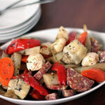 A white dish filled with a colorful antipasto salad, featuring sliced carrots, red bell peppers, marinated artichoke hearts, mushrooms, salami, and onions, garnished with herbs. Plates and utensils are in the background.