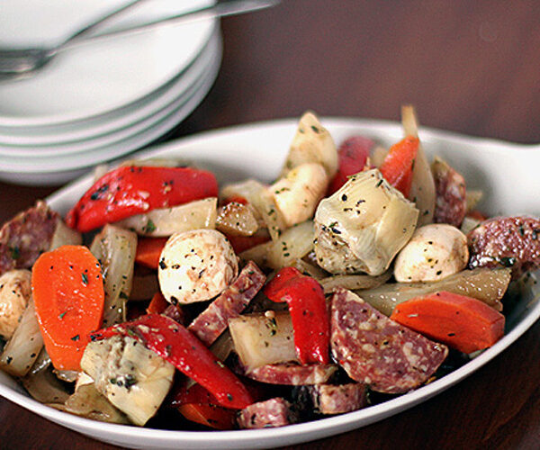 A white dish filled with a colorful antipasto salad, featuring sliced carrots, red bell peppers, marinated artichoke hearts, mushrooms, salami, and onions, garnished with herbs. Plates and utensils are in the background.