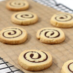 Eight round pinwheel cookies with chocolate and vanilla swirl patterns are arranged on a piece of parchment paper atop a cooling rack.