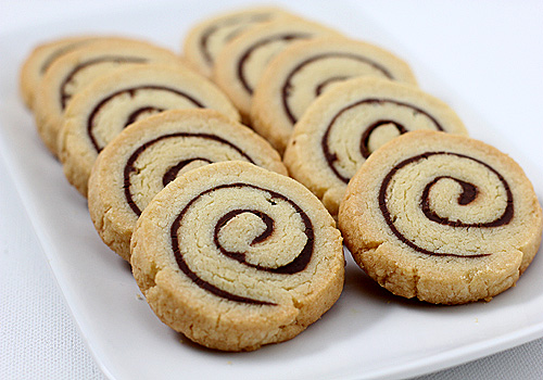 A white plate holds a row of spiral cookies with a light-colored dough and a dark chocolate swirl pattern. The cookies are neatly arranged and have a golden-brown edge.