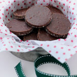 A metal bowl lined with white and pink polka dot paper holds round, chocolate cookies. A green ribbon with white stitching lies in front of the bowl.