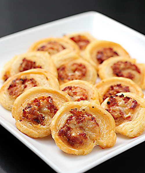 A white plate holds several golden-brown puff pastry pinwheels filled with a savory mixture, arranged neatly on a dark surface.