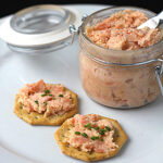 A glass jar of salmon spread with a small knife sits on a white plate next to two crackers topped with the spread and garnished with chopped chives.