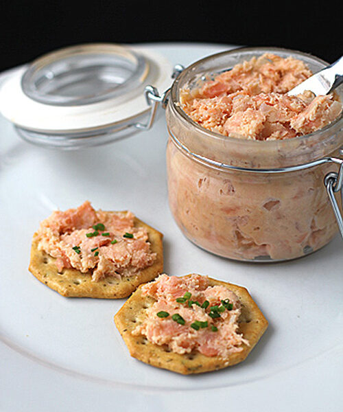A glass jar of salmon spread with a small knife sits on a white plate next to two crackers topped with the spread and garnished with chopped chives.