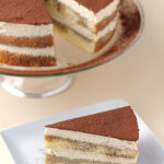 A slice of tiramisu cake with creamy layers and cocoa powder on top sits on a white plate with a fork, while the remaining cake is displayed on a glass stand in the background.