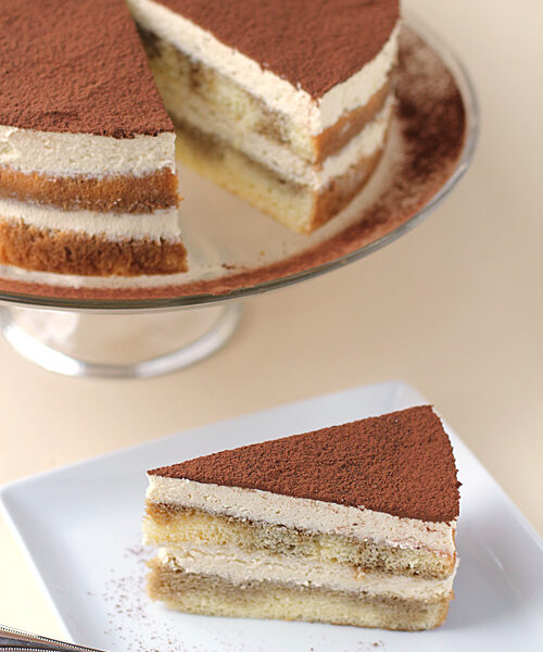 A slice of tiramisu cake with creamy layers and cocoa powder on top sits on a white plate with a fork, while the remaining cake is displayed on a glass stand in the background.