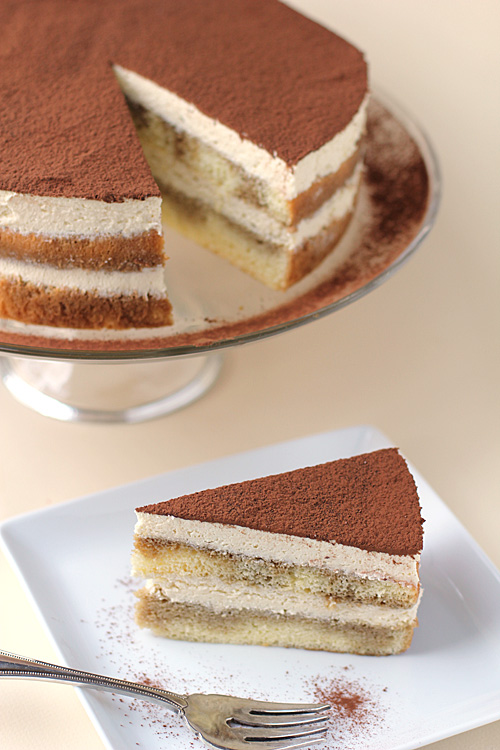 A slice of tiramisu cake with creamy layers and cocoa powder on top sits on a white plate with a fork, while the remaining cake is displayed on a glass stand in the background.