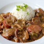 A plate of gumbo with sliced sausage, shredded meat, and vegetables in a brown sauce, served with a mound of white rice topped with chopped green onions.