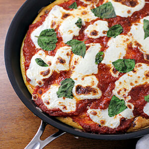 A freshly baked Margherita pizza in a black skillet, topped with melted mozzarella cheese, tomato sauce, and fresh basil leaves, sits on a wooden surface.