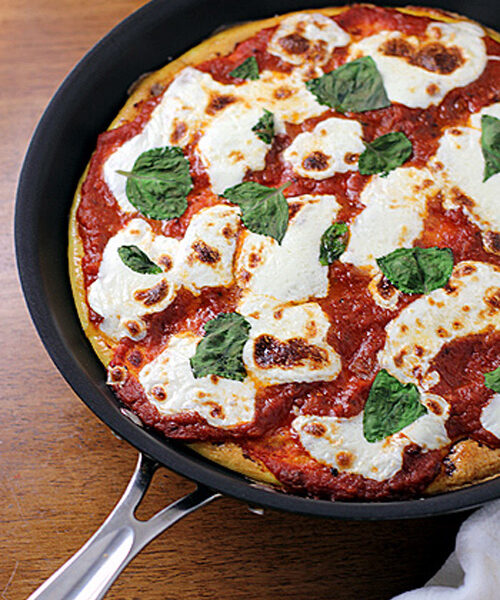 A freshly baked Margherita pizza in a black skillet, topped with melted mozzarella cheese, tomato sauce, and fresh basil leaves, sits on a wooden surface.