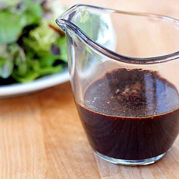 A glass pitcher filled with dark balsamic vinaigrette sits on a wooden surface, with a plate of mixed green salad slightly out of focus in the background.