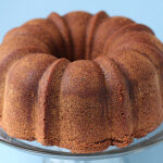 A golden-brown bundt cake sits on a glass cake stand against a light blue background. The cake has a ring shape with fluted edges and a slightly textured surface.