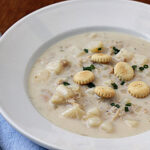 A bowl of creamy clam chowder with chunks of potatoes, garnished with chopped chives and round oyster crackers, served on a white plate next to a spoon on a blue napkin.