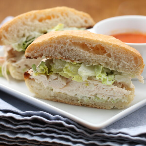 A close-up of a chicken sandwich with lettuce on ciabatta bread, cut in half and served on a white plate next to a small bowl of soup, all placed on a stack of blue napkins.