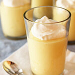 Four clear glasses filled with creamy yellow pudding, each topped with a dollop of whipped cream, are arranged on a light cloth with a spoon nearby.