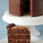 A slice of German chocolate cake with chocolate frosting and coconut-pecan filling sits on a white plate in front of the remaining cake on a stand. A fork rests beside the slice.