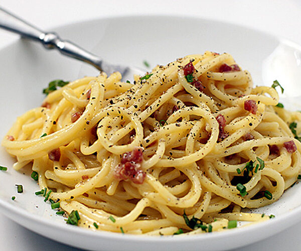 A plate of spaghetti carbonara topped with finely chopped parsley, crispy bacon bits, and a sprinkle of black pepper, with a fork resting on the side of the white bowl.