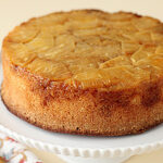 A round pineapple upside-down cake sits on a white cake stand, showing caramelized pineapple slices on top and a golden brown edge. A patterned napkin and fork are partially visible beside the stand.