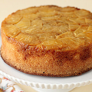 A round pineapple upside-down cake sits on a white cake stand, showing caramelized pineapple slices on top and a golden brown edge. A patterned napkin and fork are partially visible beside the stand.