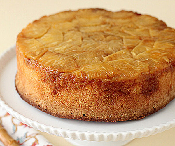 A round pineapple upside-down cake sits on a white cake stand, showing caramelized pineapple slices on top and a golden brown edge. A patterned napkin and fork are partially visible beside the stand.