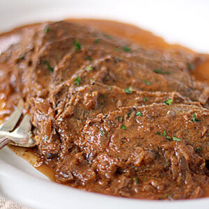 Slices of tender beef covered in a rich brown gravy with herbs, served on a white plate with a fork beside the meat.