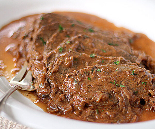 Slices of tender beef covered in a rich brown gravy with herbs, served on a white plate with a fork beside the meat.