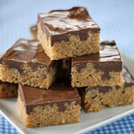 A plate stacked with chocolate-topped crispy rice treats sits on a blue and white checkered tablecloth. The bars have a thick chocolate layer on top and a golden, textured base.