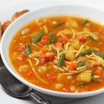 A bowl of vegetable noodle soup with green beans, carrots, potatoes, and noodles in a tomato broth, served with two slices of bread on a white plate and a spoon beside the bowl.