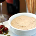 A white bowl filled with smooth pâté sits on a plate with caper berries and pickled peppers, with a glass of red wine, a bottle, and a vase of breadsticks blurred in the background.
