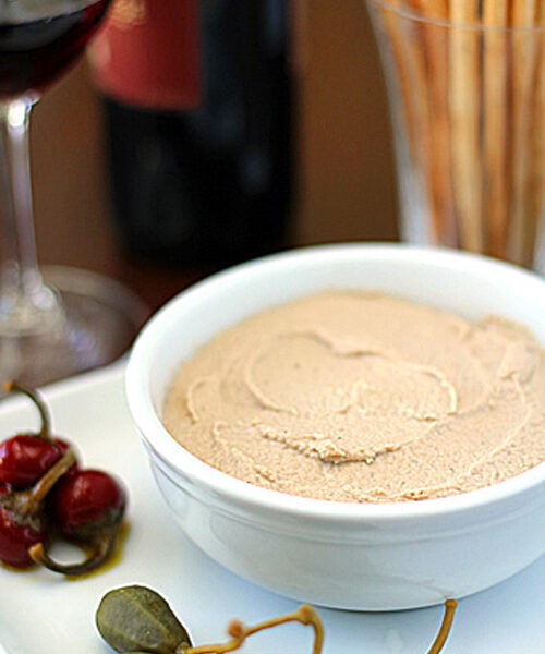 A white bowl filled with smooth pâté sits on a plate with caper berries and pickled peppers, with a glass of red wine, a bottle, and a vase of breadsticks blurred in the background.