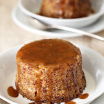 Two individual sticky toffee puddings sit on white plates, topped with rich caramel sauce that drips down the sides. Each dessert is accompanied by a spoon, with a light creamy sauce pooling at the base.