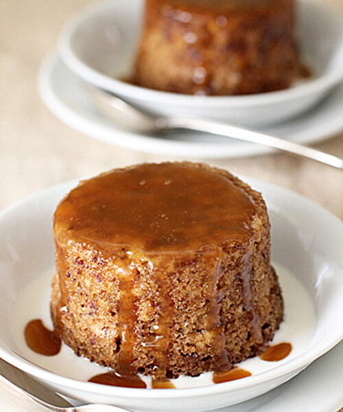 Two individual sticky toffee puddings sit on white plates, topped with rich caramel sauce that drips down the sides. Each dessert is accompanied by a spoon, with a light creamy sauce pooling at the base.