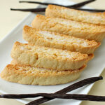 Five pieces of vanilla biscotti are arranged on a rectangular white plate, garnished with three whole vanilla beans placed around them.