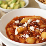 A white bowl filled with a stew containing potatoes, beans, and sausage in a red sauce, topped with crumbled cheese. In the background, there are bowls of diced avocado and tortilla chips.