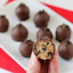 A hand holds a chocolate-coated ball with a bite taken out, revealing a cookie dough center and chocolate chips. More chocolate balls are arranged on a white plate in the background.