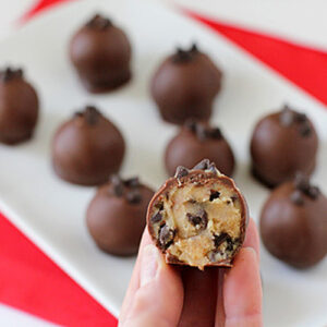 A hand holds a chocolate-coated ball with a bite taken out, revealing a cookie dough center and chocolate chips. More chocolate balls are arranged on a white plate in the background.
