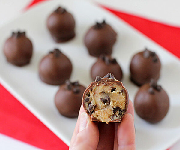A hand holds a chocolate-coated ball with a bite taken out, revealing a cookie dough center and chocolate chips. More chocolate balls are arranged on a white plate in the background.