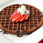 A round chocolate waffle topped with sliced strawberries, whipped cream, and chocolate chips, drizzled with chocolate sauce, served on a white square plate with a fork nearby.