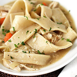 A bowl of homemade chicken and noodles with wide egg noodles, shredded chicken, carrots, and parsley in a savory broth.