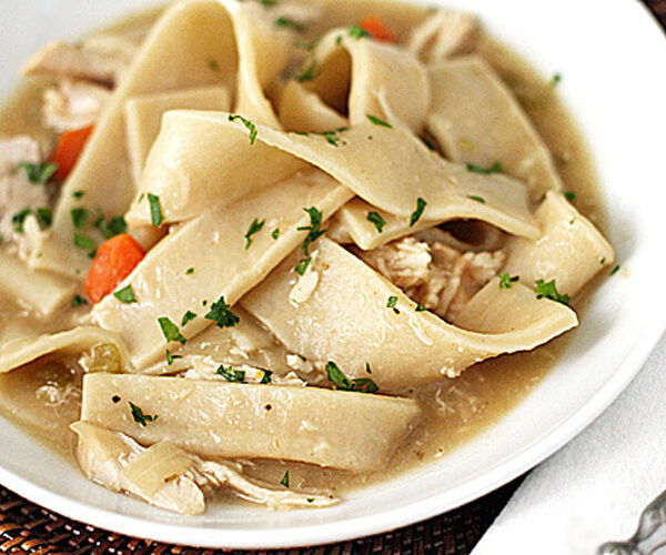 A bowl of homemade chicken and noodles with wide egg noodles, shredded chicken, carrots, and parsley in a savory broth.