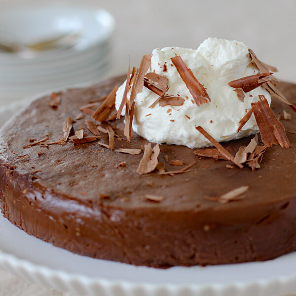 A chocolate cake topped with a dollop of whipped cream and chocolate shavings sits on a white plate, with a blurred stack of plates and spoons in the background.