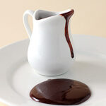 A small white pitcher with chocolate sauce dripping down its side sits on a white plate, with a puddle of chocolate sauce on the plate in front of it.